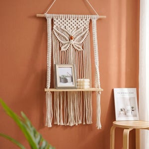 May include: Cream-colored macrame wall hanging with a wooden shelf. The macrame features a geometric design and a wooden dowel at the top. A framed photo and a candle sit on the shelf. The Minimal Home book is on a small wooden table.