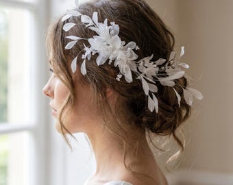 Könnte beinhalten: Ein weißes Blumenschmuck-Haarteil mit zarten Federn und Perlenakzenten. Das Haarteil wird seitlich am Kopf getragen, wobei die Federn und Blüten nach unten fallen. Geeignet für Hochzeiten oder besondere Anlässe.