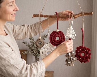 Könnte beinhalten: Ein Wandbehang aus einem natürlichen Holzast, an dem fünf Makramee-Ornamente hängen. Die Ornamente umfassen kreisförmige Designs in Rot, Weiß und Beige sowie ein baumförmiges Design in Weiß mit beigen Fransen und kleinen weißen Perlen.