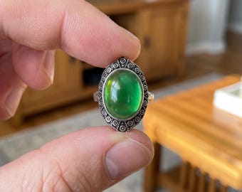 Könnte beinhalten: Ein silberfarbener Ring mit einem großen, glatten, ovalen grünen Stimmungsstein, der in einen dekorativen Metallrahmen mit Perlenrand gefasst ist. Die Fassung hat eine spitze, marquiseartige Form mit detaillierten kreisförmigen Verzierungen.