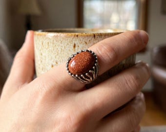 Könnte beinhalten: Ein Ring mit einem ovalen, rotbraunen Goldfluss-Cabochon mit funkelnden Einschlüssen. Der Stein ist in eine silberfarbene Metallschiene mit einer dekorativen, gezackten Fassung eingefasst, die nach oben ragt und die Basis des Steins umrahmt.