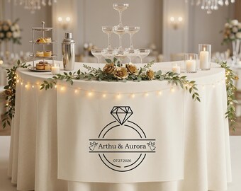 Könnte beinhalten: Ein Hochzeitsempfangstisch mit cremefarbener Tischdecke, verziert mit Lichterketten und Blumengestecken. Ein Champagnerturm und Gläser sind aufgebaut, zusammen mit einem mehrstöckigen Dessertständer und einem Cocktailshaker. Der Tischrock zeigt eine Diamantring-Grafik mit den Namen "Arthu & Aurora" und dem Datum 07.27.2026.