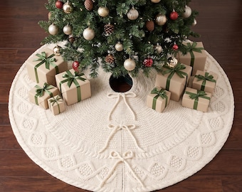 Könnte beinhalten: Ein runder, cremefarbener Strick-Weihnachtsbaumteppich mit einem strukturierten Zopfstrickmuster am Rand und drei dekorativen Bindebändern in der Mitte. Der Baumteppich liegt unter einem geschmückten Weihnachtsbaum mit Geschenken.