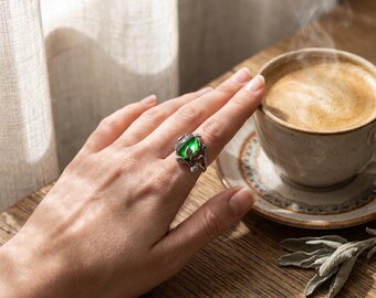 Könnte beinhalten: Ein Silberring mit einem polierten, ovalen grünen Edelstein, der in ein detailliertes Metallband mit kleinen Blättern und Ranken eingefasst ist. Der Ring wird an einem Finger getragen und zeigt den leuchtend grünen Stein sowie die filigrane Arbeit.