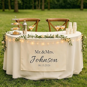 May include: A wedding reception table with a cream-colored tablecloth, adorned with string lights and floral garlands. The table is set with cocktails, desserts, and a sign that reads "Mr.&Mrs. Johnson 06.15.2026". Two wooden chairs are in the background.