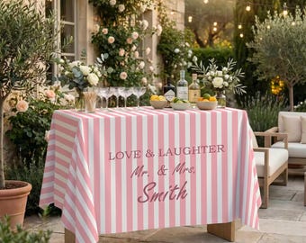 Könnte beinhalten: Ein rechteckiger Tisch mit einer rosa-weiß gestreiften Tischdecke, bedruckt mit dem Text LOVE & LAUGHTER Mr. & Mrs. Smith. Der Tisch ist mit klaren Gläsern, Spirituosenflaschen und Schalen mit Zitrusfrüchten in einer Außenumgebung gedeckt.