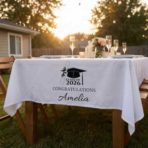 Puede incluir: Un mantel blanco con un diseño de graduación. El diseño incluye un birrete, un diploma y el texto "Class of 2026 Congratulations Amelia" en negro. La mesa está puesta al aire libre con copas de champán.