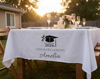 Könnte beinhalten: Ein weißes Tischtuch mit einem Abschluss-Design. Das Design umfasst eine Abschlusskappe, ein Diplom und den Text "Class of 2026 Congratulations Amelia" in Schwarz. Der Tisch ist im Freien mit Sektgläsern gedeckt.