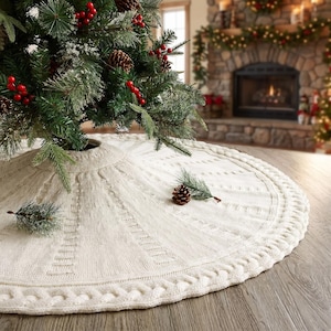 May include: A circular, off-white knitted tree skirt featuring a textured cable knit pattern and a braided border. The skirt is positioned beneath a green Christmas tree decorated with red berries and pinecones, creating a festive holiday display.