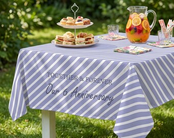 Könnte beinhalten: Eine hellviolett-weiß diagonal gestreifte Tischdecke mit dem aufgedruckten Text TOGETHER & FOREVER Our 6 Anniversary. Die Tischdecke liegt auf einem Tisch, der mit einer zweistöckigen Etagere, einer Glaskaraffe mit Fruchtpunsch und Gläsern gedeckt ist.