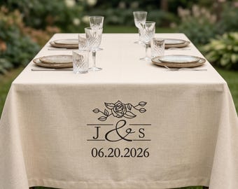 Könnte beinhalten: Eine beige Leinentischdecke für eine formelle Outdoor-Veranstaltung. Die Tischdecke ist mit einem Blumendesign, Initialen und dem Datum 06.20.2026 verziert. Der Tisch ist mit Tellern, Gläsern und Besteck gedeckt.