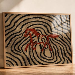 May include: A framed rectangular wall art print featuring a stylized red horse illustration centered against a background of concentric, wavy black lines on a tan surface. The artwork is enclosed in a simple, light-toned wooden frame.