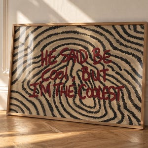 Puede incluir: Una pieza de arte mural rectangular enmarcada con fondo beige y líneas negras concéntricas garabateadas. El texto HE SAID BE COOL, BUT I'M THE COOLEST está impreso en un estilo de letra manual, negrita y color rojo oscuro.
