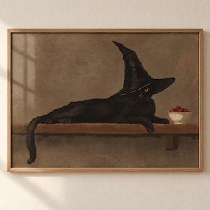 May include: A framed wall art print depicting a black cat wearing a pointed black witch hat, lying on a wooden shelf next to a small white bowl filled with red cherries. The artwork features a muted brown background and a light wood frame.