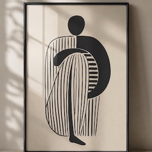 Puede incluir: Impresión artística abstracta en blanco y negro en un marco negro. La obra presenta un diseño minimalista de una figura con una prenda a rayas y una cabeza y un brazo negros. El fondo es de color beige neutro.