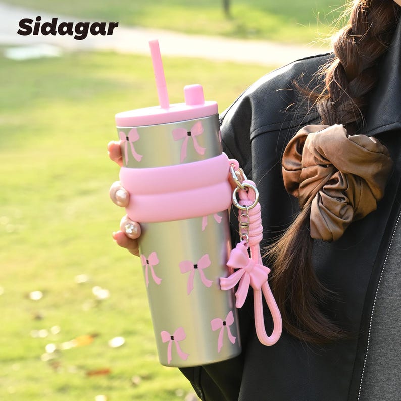 May include: A silver stainless steel tumbler featuring a repeating pattern of small pink bows. The tumbler has a pink silicone lid with a matching straw, a pink silicone sleeve around the middle, and a pink bow charm attached to a wrist strap.