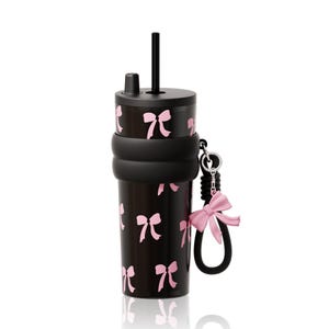 May include: A black tumbler featuring a repeating pattern of light pink bows. The cup includes a black lid with a straw, a thick black silicone boot around the middle, and a black wrist strap attached to a silver ring with a pink bow charm.