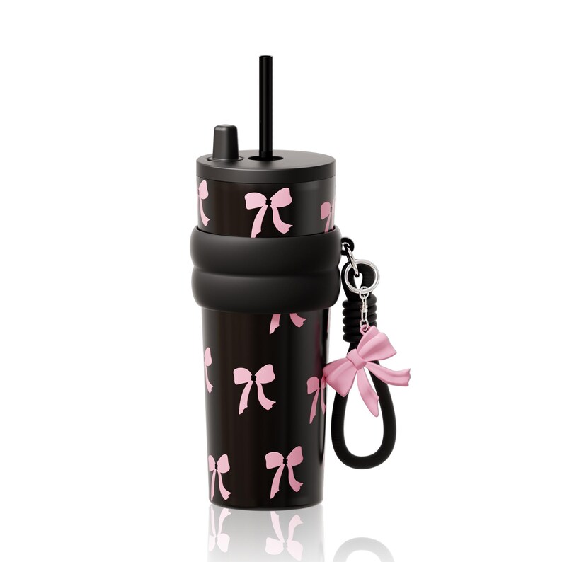 May include: A black tumbler featuring a repeating pattern of light pink bows. The cup includes a black lid with a straw, a thick black silicone boot around the middle, and a black wrist strap attached to a silver ring with a pink bow charm.