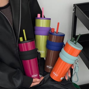 May include: Four cylindrical tumblers with lids and straws, each featuring a silicone grip band and a carrying strap. The tumblers are magenta, lime green, brown, and orange, all displaying the white Sidagar logo on the lower front section.