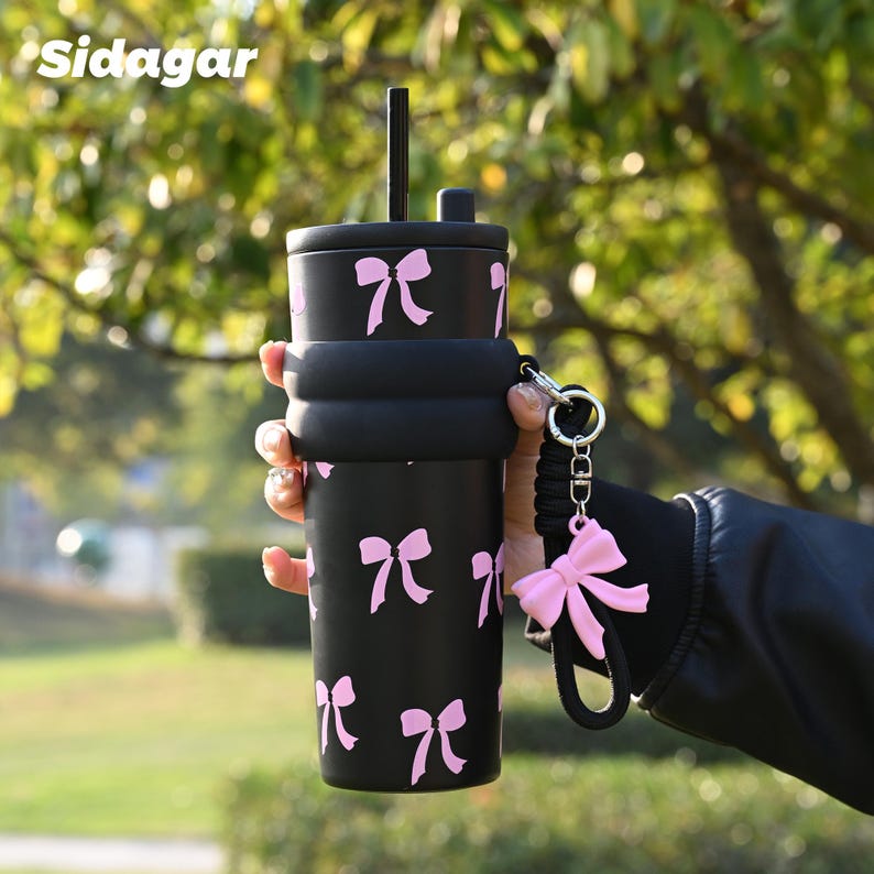 May include: A matte black tumbler featuring a repeating pattern of light pink bows, a black lid with a matching straw, a thick black silicone sleeve around the center, and a detachable black wrist strap with a large pink bow charm. Text reads Sidagar.