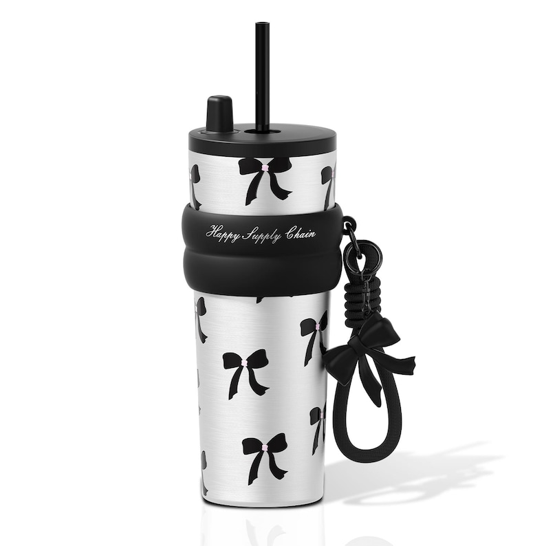 May include: A silver stainless steel tumbler featuring a black bow pattern, a black lid with a straw, and a black silicone sleeve printed with the text Happy Supply Chain. A black silicone strap with a decorative bow charm is attached to the side.