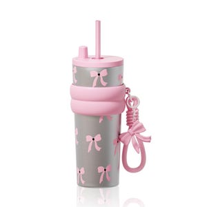 May include: A silver-toned tumbler featuring a repeating pattern of small pink bows. The cup includes a pink lid with a matching straw, a thick pink silicone band around the center, and a pink bow-shaped charm attached to the side by a ring.
