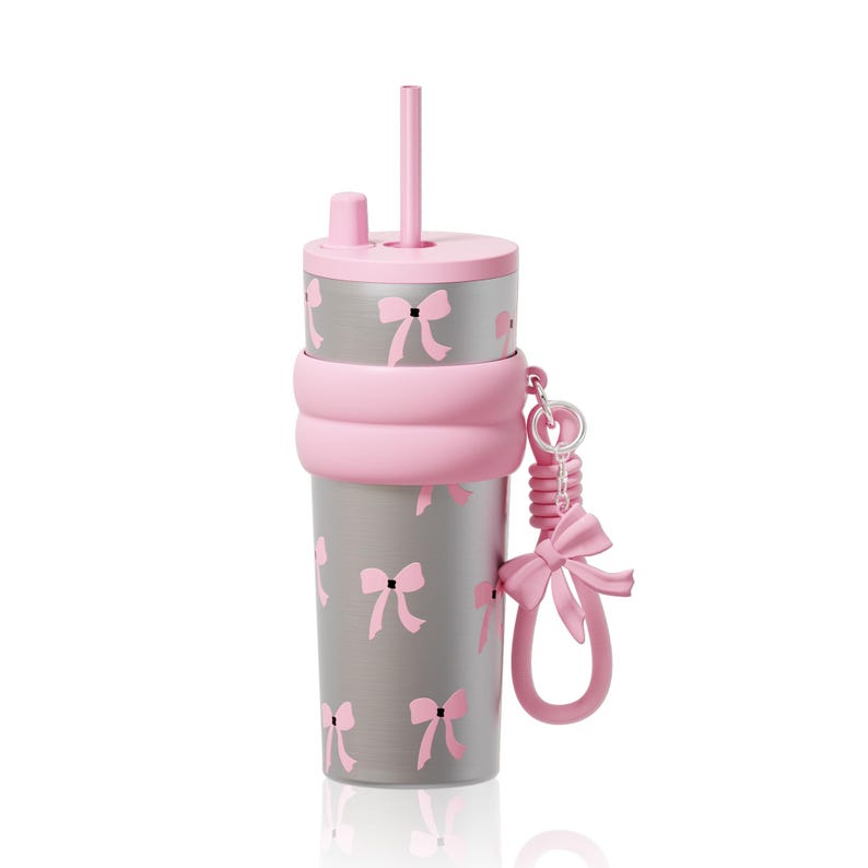 May include: A silver-toned tumbler featuring a repeating pattern of small pink bows. The cup includes a pink lid with a matching straw, a thick pink silicone band around the center, and a pink bow-shaped charm attached to the side by a ring.
