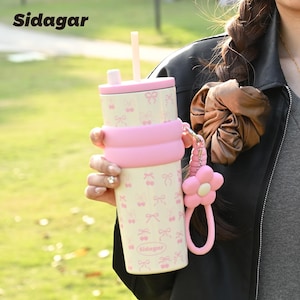 May include: A cream-colored tumbler with a repeating pink bow pattern, featuring a pink silicone sleeve, a matching lid with a straw, and a pink flower-shaped charm attached to the side. The brand name Sidagar is printed on the tumbler.