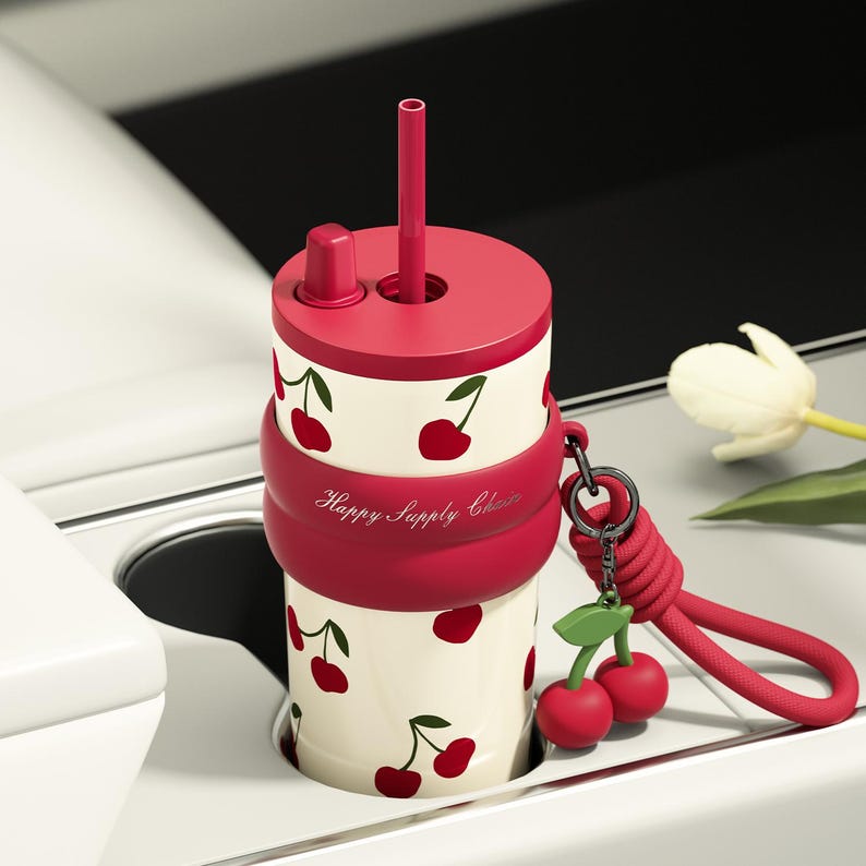 May include: A cylindrical tumbler with a cream-colored body featuring a red cherry pattern. It has a red lid with a straw and a red silicone band embossed with the text Happy Supply Chain. A red braided lanyard with a cherry charm is attached.
