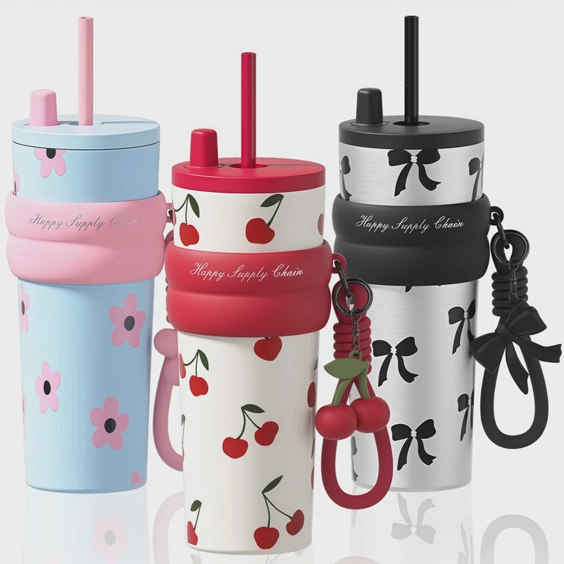 May include: Three cylindrical tumblers with lids and straws. The left is light blue with pink flowers, the center is white with red cherries, and the right is silver with black bows. Each features a silicone band labeled Happy Supply Chain.