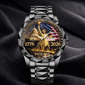 May include: A black wristwatch with a detailed face featuring the Statue of Liberty, the U.S. Capitol, and an American flag. The watch face includes the text "UNITED STATES OF AMERICA", "1776", and "2026". The watch band is a matching black metal.