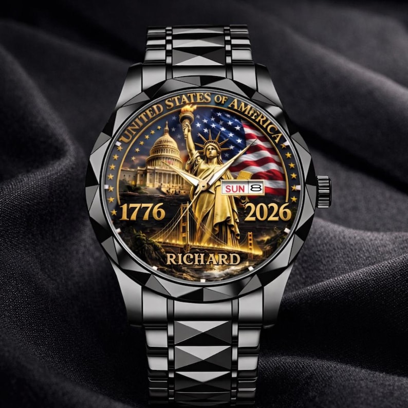 May include: A black wristwatch with a detailed face featuring the Statue of Liberty, the U.S. Capitol, and an American flag. The watch face includes the text "UNITED STATES OF AMERICA", "1776", and "2026". The watch band is a matching black metal.