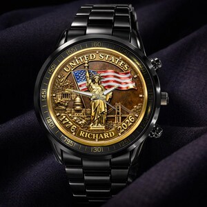 May include: Black wristwatch with a gold-toned face featuring the Statue of Liberty, an American flag, and the text "UNITED STATES 1776 RICHARD 2026". The watch has a black metal band and a gold-toned bezel with minute markers.