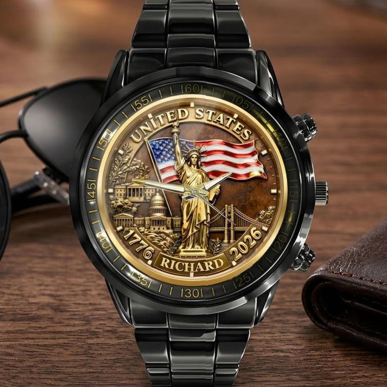 May include: Black wristwatch with a detailed face featuring the Statue of Liberty, an American flag, and the text "UNITED STATES", "1776", and "2026". The watch has a black metal band and a brown face.