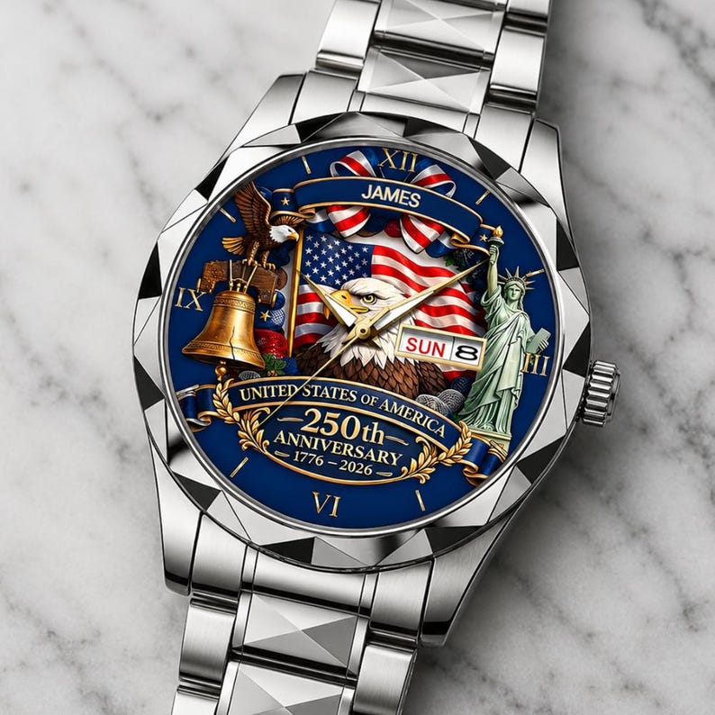 May include: A silver-tone analog wristwatch with a blue dial featuring a bald eagle, the Statue of Liberty, a Liberty Bell, and an American flag. The dial displays the text JAMES, UNITED STATES OF AMERICA, 250th ANNIVERSARY, 1776-2026, and SUN 8.