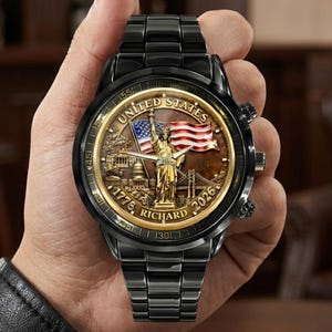 May include: Black wristwatch with a detailed face featuring the Statue of Liberty, the American flag, and the text "UNITED STATES" and "RICHARD". The watch has a black metal band and a gold-colored bezel.