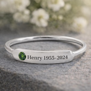 May include: A silver-colored ring with a green gemstone and the engraved text "Henry 1955-2024". The ring is set on a gray stone surface, with blurred white flowers in the background.