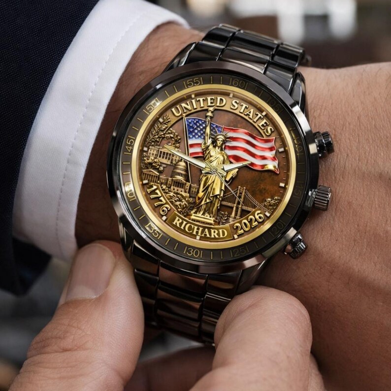 May include: A black wristwatch with a gold-toned face featuring the Statue of Liberty, an American flag, and the text "UNITED STATES", "1776", "RICHARD", and "2026". The watch has a black metal band.