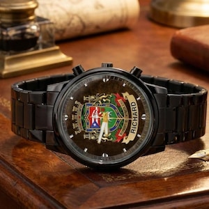 May include: Black wristwatch with a round face featuring a colorful emblem and the name "Richard" on a banner. The watch has a black metal band and a dark brown face. The watch is displayed on a wooden surface.