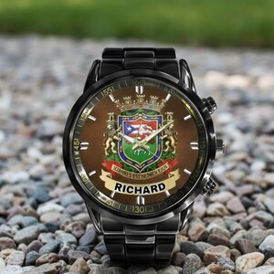 May include: A black wristwatch with a brown face featuring a coat of arms design. The watch face includes the name "RICHARD" and the text "JOANNES EST NOMEN EIUS". The watch has a black metal band and is set against a background of small stones.