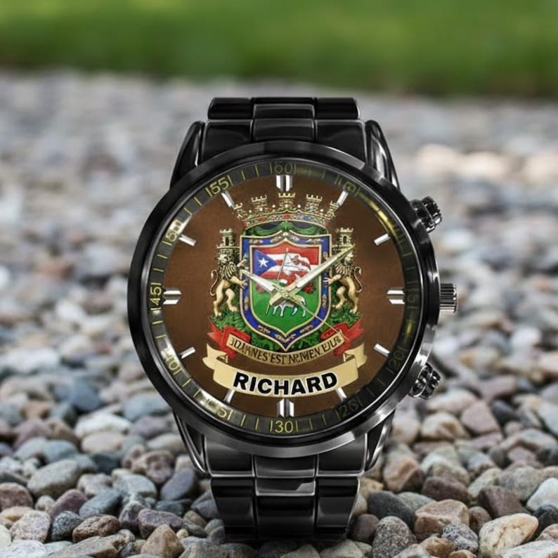May include: A black wristwatch with a brown face featuring a coat of arms design. The watch face includes the name "RICHARD" and the text "JOANNES EST NOMEN EIUS". The watch has a black metal band and is set against a background of small stones.