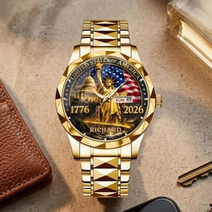 May include: Gold-coloured wristwatch with a detailed face depicting the Statue of Liberty, the American flag, and the Capitol building. The watch face includes the text "UNITED STATES OF AMERICA", "1776", "2026", and "RICHARD". The watch band has a geometric design.