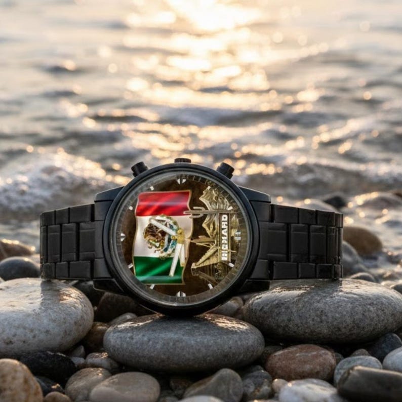 May include: Black wristwatch with a Mexican flag design on the face. The watch has a black metal band and is resting on a bed of smooth, gray stones near the water. The watch face also has the name "RICHARD" on it.