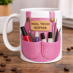 May include: White ceramic mug with a pink apron design, featuring the text "NAIL TECH SOPHIA". The apron pockets hold nail polish bottles, tools, and glitter. Gold-coloured buttons accent the apron design.