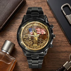 May include: Black wristwatch with a gold-toned face featuring the Statue of Liberty, an American flag, and the text "United States 1776-2026". The watch has a black metal band and a detailed design.