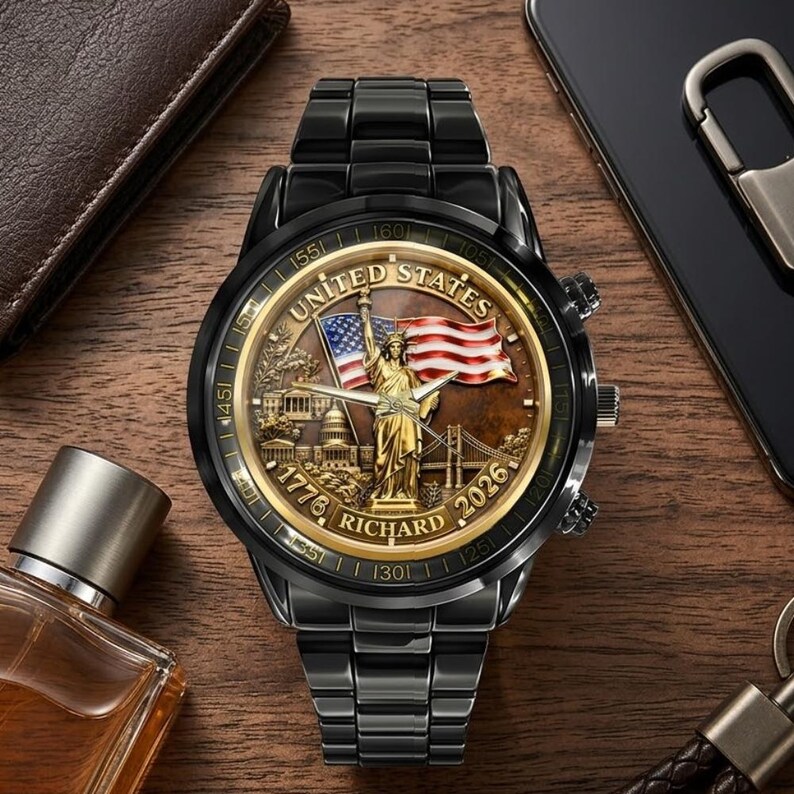May include: Black wristwatch with a gold-toned face featuring the Statue of Liberty, an American flag, and the text "United States 1776-2026". The watch has a black metal band and a detailed design.