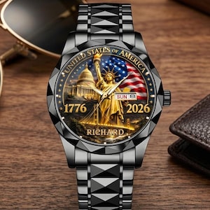 May include: A black wristwatch with a detailed face featuring the Statue of Liberty, the American flag, and the U.S. Capitol. The watch face also displays the text "UNITED STATES OF AMERICA", "1776", "2026", and "RICHARD". The watch has a black metal band.