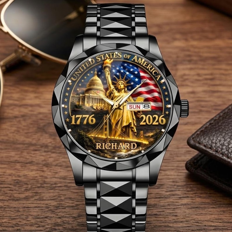 May include: A black wristwatch with a detailed face featuring the Statue of Liberty, the American flag, and the U.S. Capitol. The watch face also displays the text "UNITED STATES OF AMERICA", "1776", "2026", and "RICHARD". The watch has a black metal band.