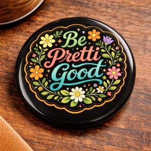 May include: A black round button with the words "Be Pretti Good" in colorful script. The text is surrounded by a floral design with yellow, orange, pink, and purple flowers and green leaves. The button is on a wooden surface.