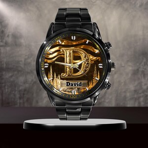 K&ouml;nnte beinhalten: Schwarze Armbanduhr mit goldenem Zifferblatt, das den Buchstaben "D" und den Namen "David" zeigt. Die Uhr hat ein schwarzes Metallarmband und ein rundes Zifferblatt mit goldenen Akzenten und einer Waldszene.